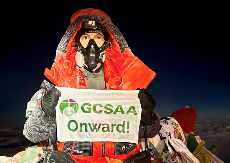Rhett Evans in cold-weather climbing gear on Mount Everest, holding a GCSAA flag