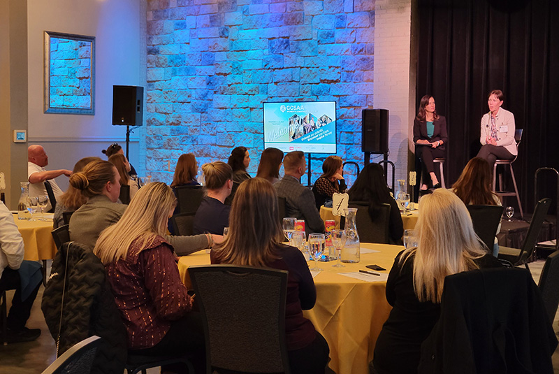Two women sitting on a stage and speaking to a large assembled group