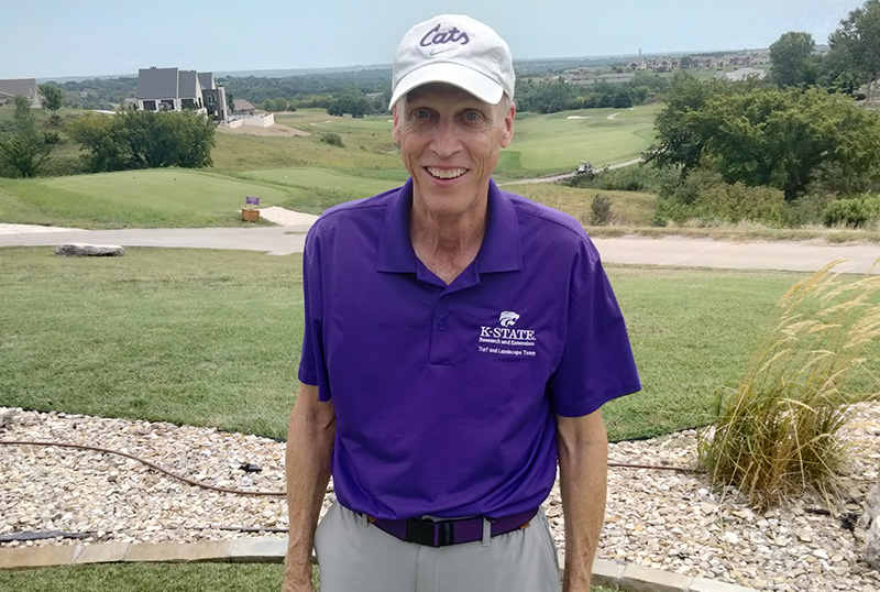 A man in a purple polo and white ball cap stands outside against a background of green turfgrass