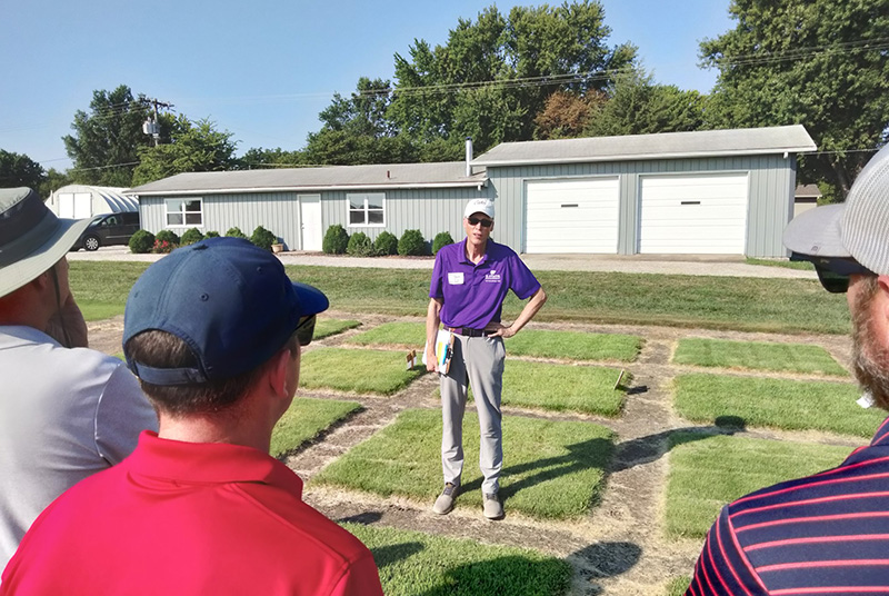 A man in a purple polo and white ball cap stands outside against a background of green turfgrass