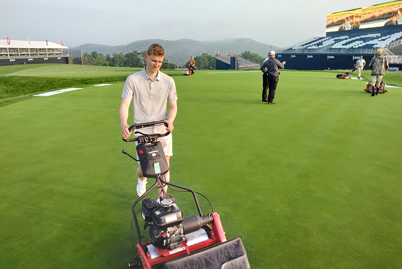 Man mowing greens on a golf course with championship stands in the background